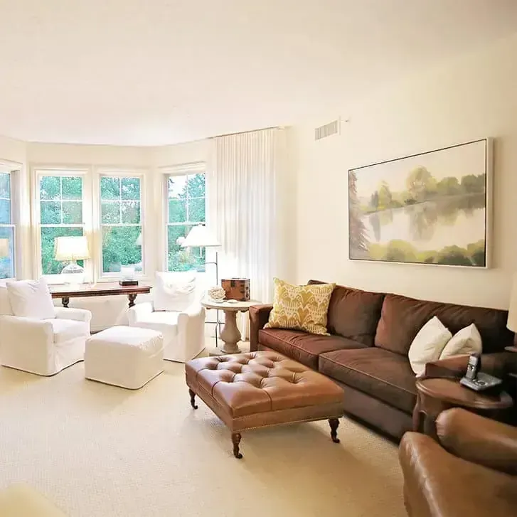 Bright living room with brown sofa, white chairs, ottoman, and large windows with sheer curtains