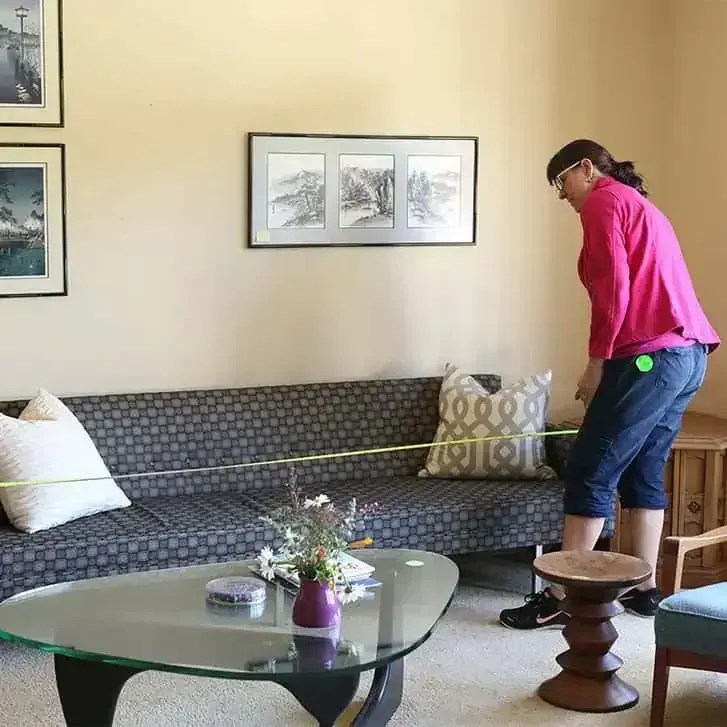 Person vacuuming a gray couch in a bright living room with a glass coffee table.