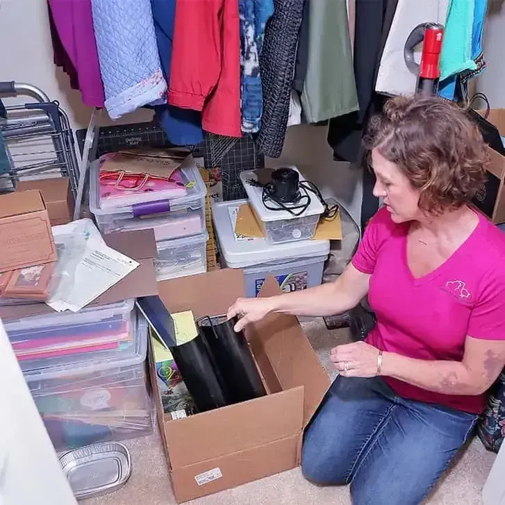 Woman in pink shirt sorting items into boxes in a cluttered room with hanging clothes.