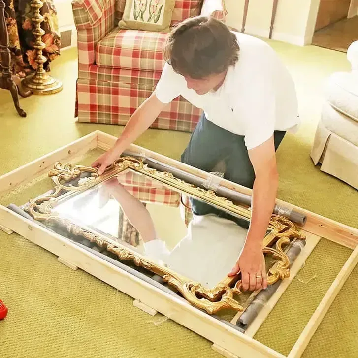 Person assembling a large framed mirror on a carpeted floor in a bright living room