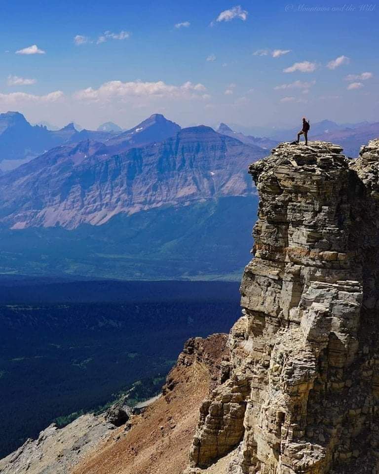 Person standing on a rocky cliff overlooking a blue mountain valley under a clear sky
