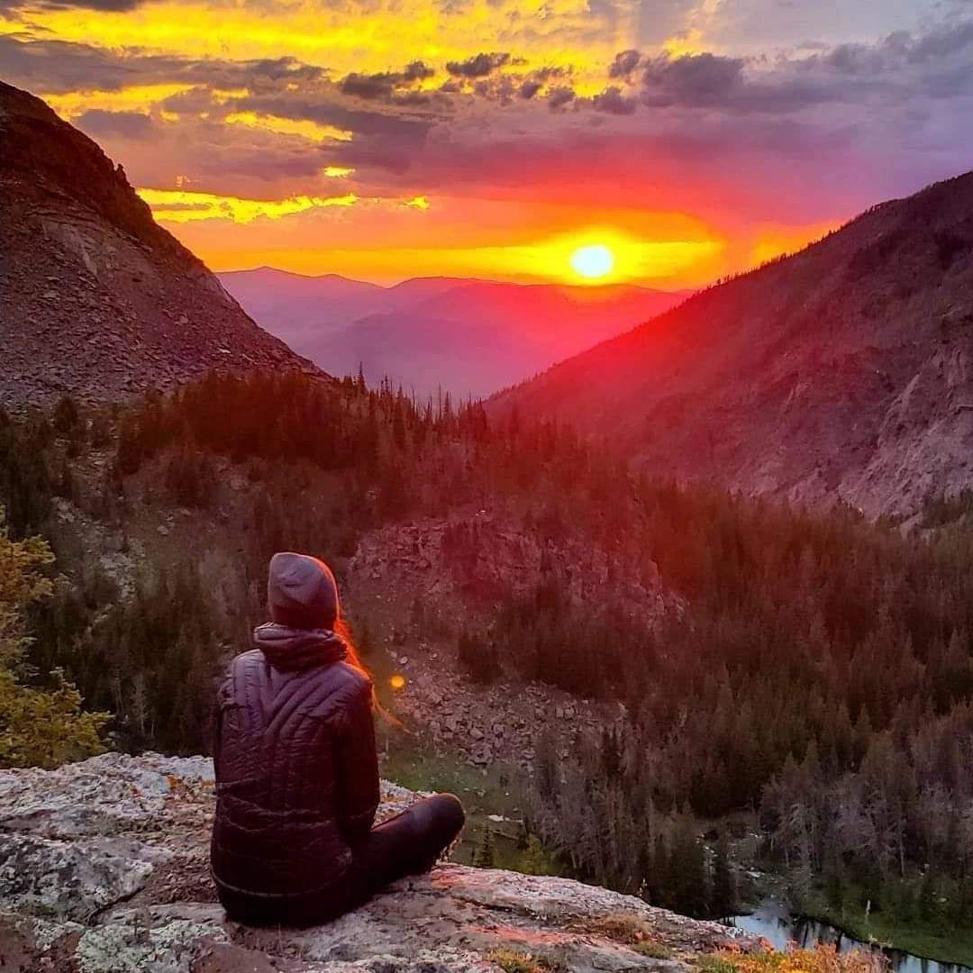 Person sitting on a rock overlooking a mountain valley at sunset, with vivid orange and pink sky