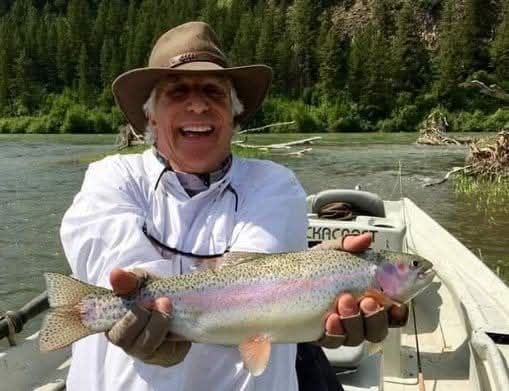 Angler in a boat holds a rainbow trout on a river with forested banks.