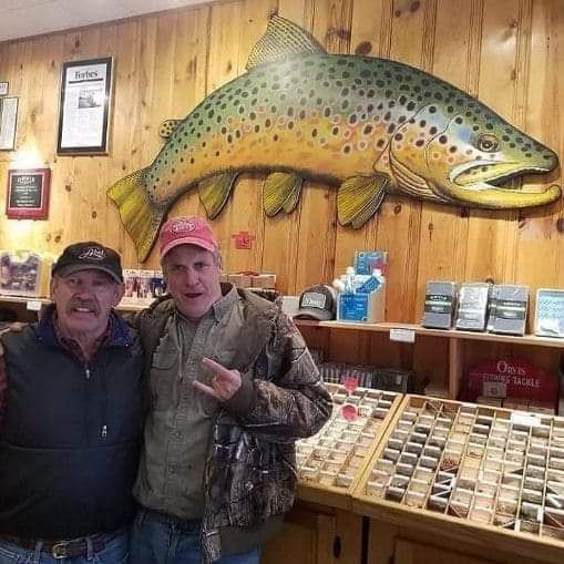 Two men posing indoors under a large mounted fish, with wooden walls and display shelves behind them