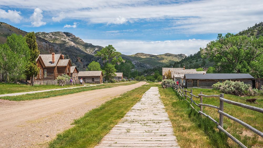 Wooden boardwalk along a dirt road in a mountain village with cabins, green hills, and blue sky.