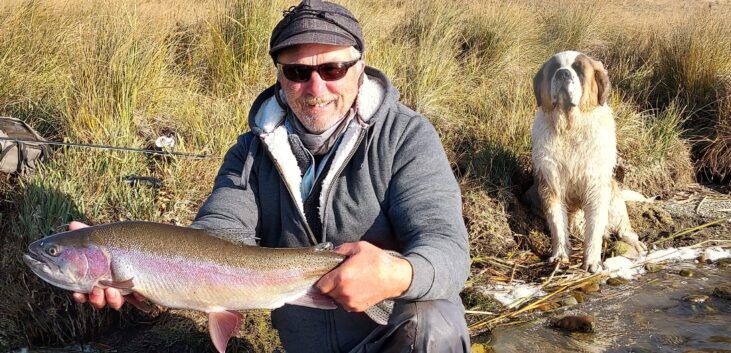 Person holding a large fish by a stream with a dog in the background