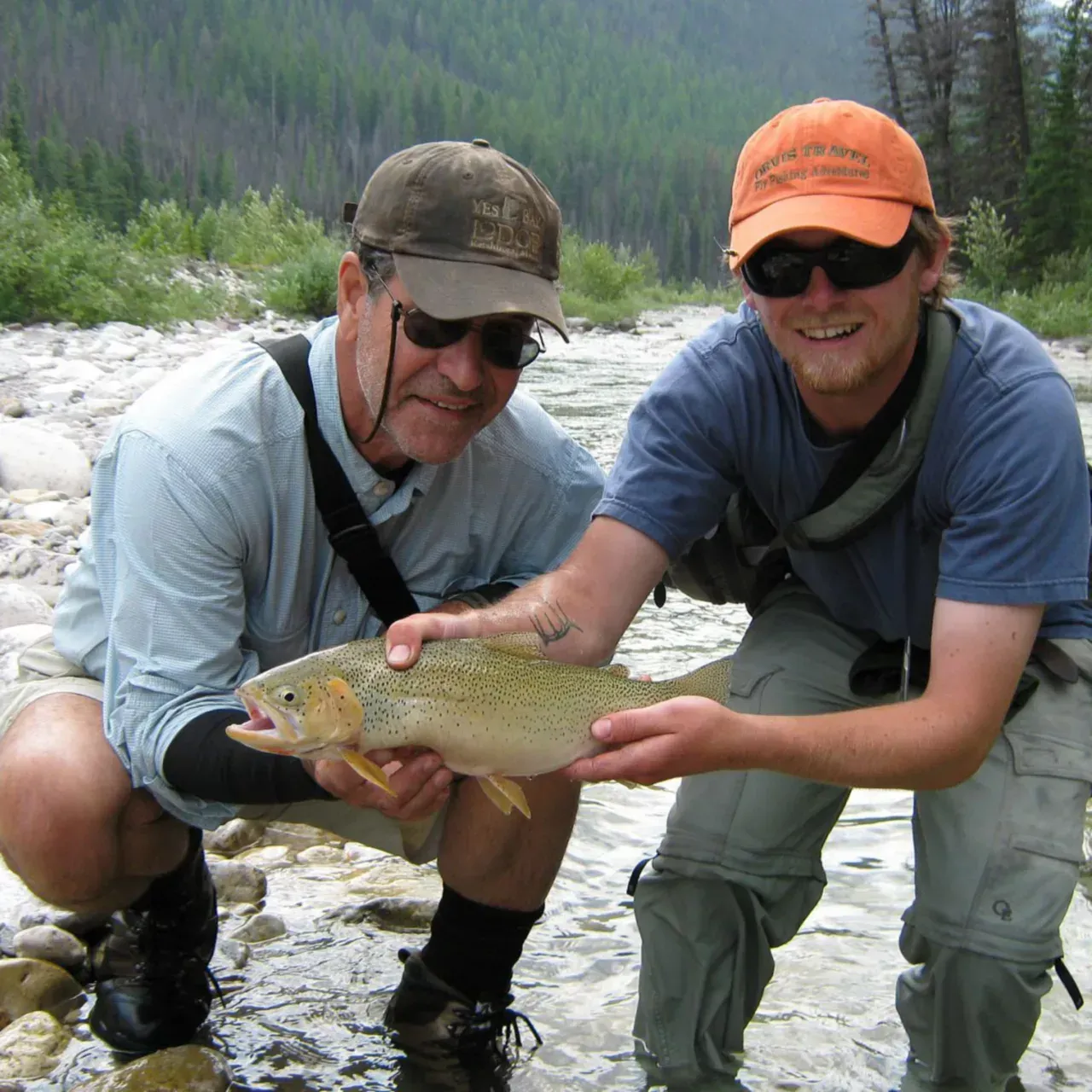 Two anglers kneel by a river, smiling and holding a fish together.