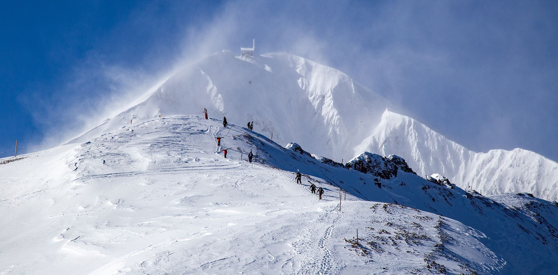 Snow-covered mountain ridge under a blue sky, with a few hikers near the summit