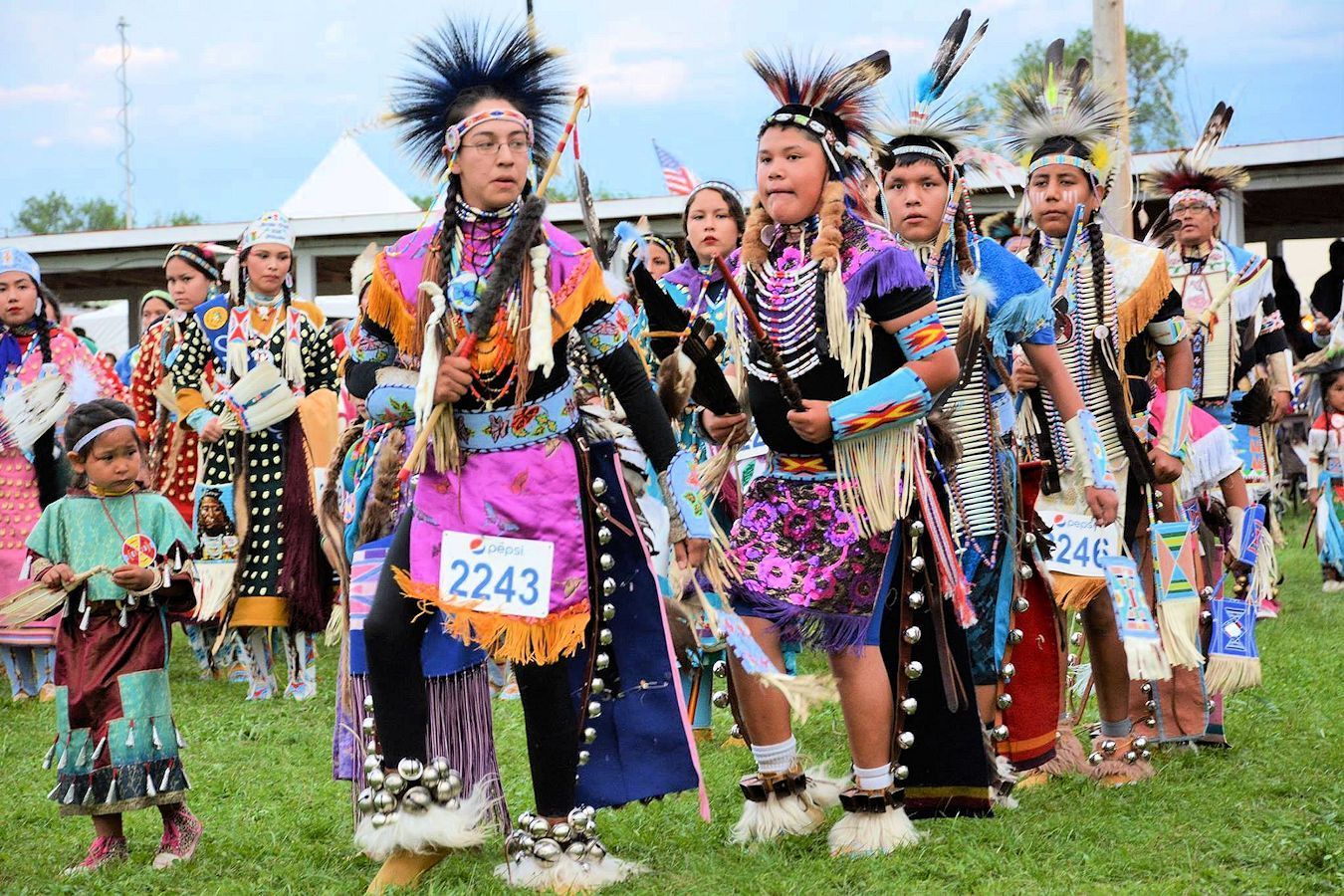 Dancers in colorful traditional regalia performing outdoors on grass, some wearing numbered bibs