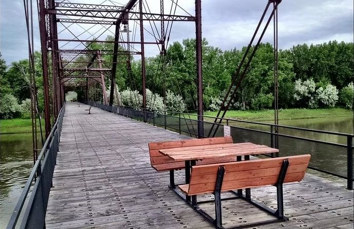 Wooden bench on a weathered footbridge over water, leading toward a tree-lined park.