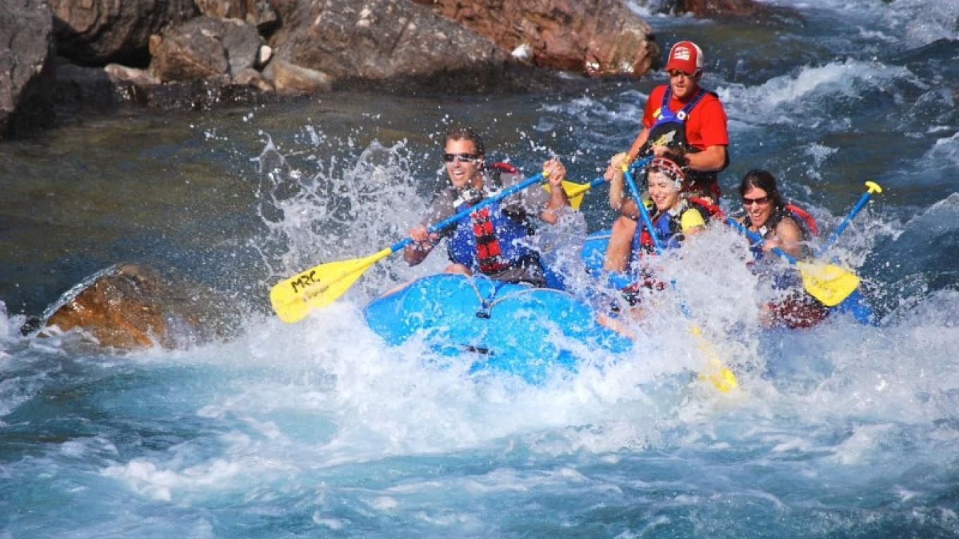 Group rafting through rough blue water, paddles raised, with a guide in a red helmet behind them.