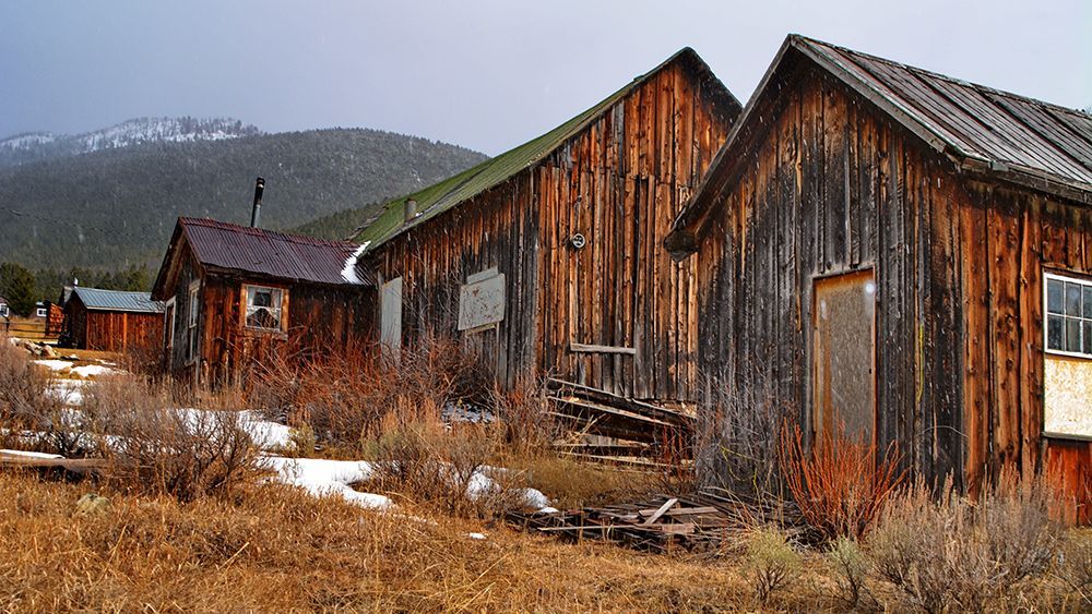 Weathered wooden cabins in a snowy, rustic mountain landscape under an overcast sky