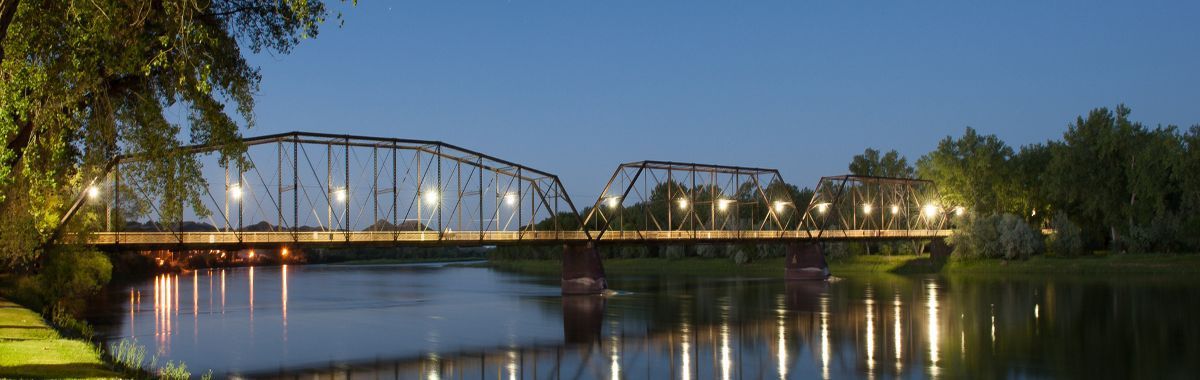 Illuminated steel bridge spanning a calm river at dusk with tree reflections and city lights