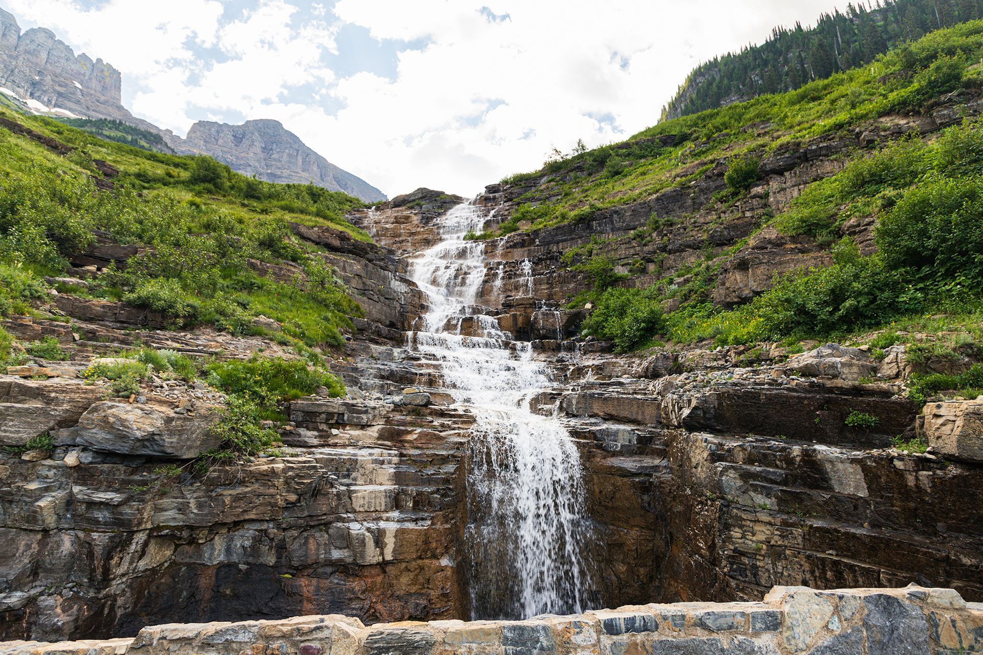 Small waterfall cascading over rocky cliffs amid green hills under a cloudy sky