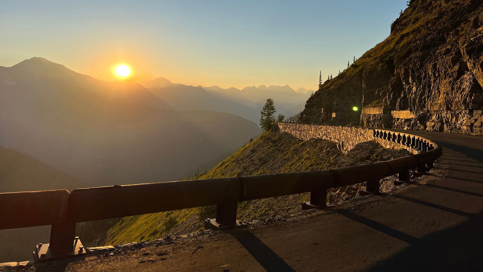 Sunset over mountain valley beside a winding road and wooden guardrail, with rocky slope and distant peaks