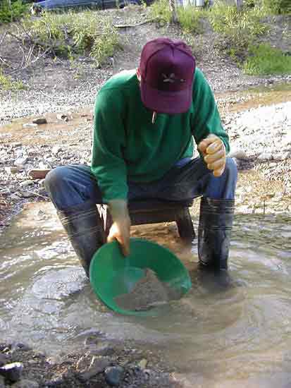 Person crouching in a stream panning for gold with a green pan and boots.