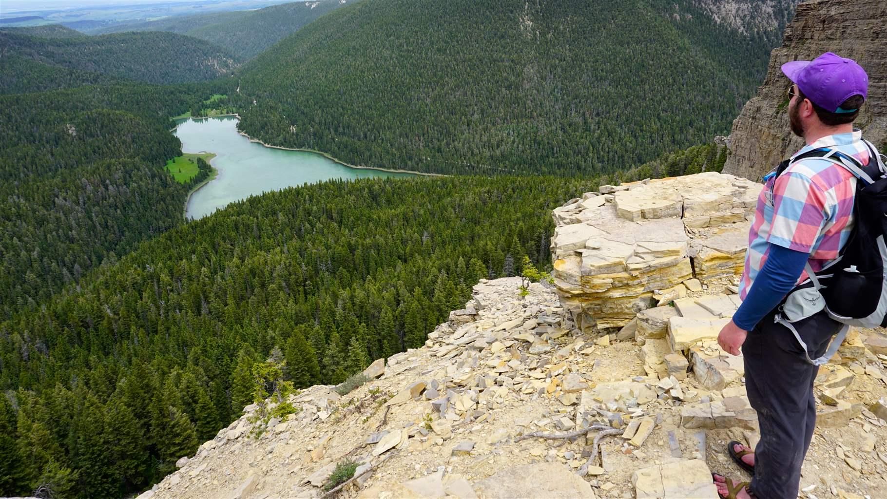 Hiker in a purple cap looks over a rocky cliff at a winding river through a green forested valley.