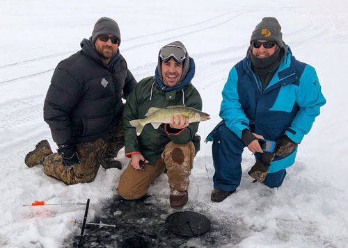 Three anglers kneel on snowy ice, holding a caught fish near an ice-fishing hole.
