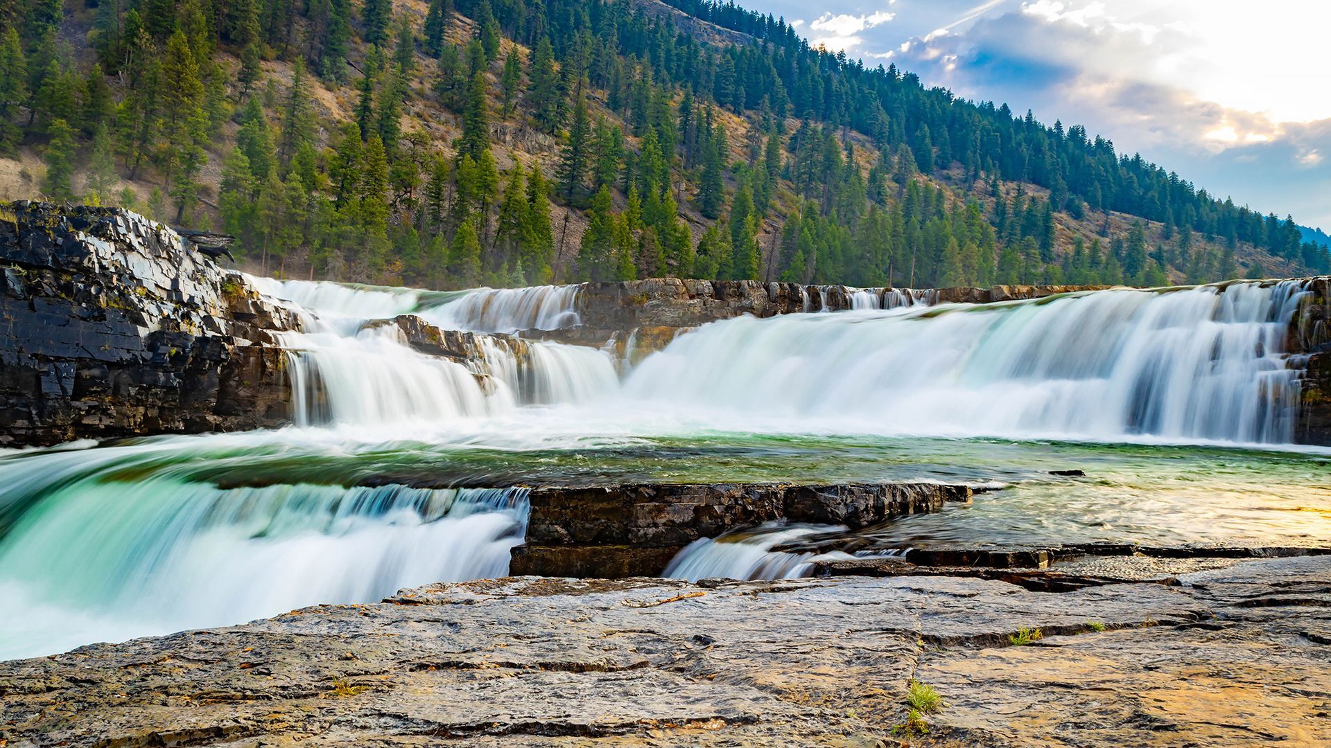 Wide waterfall cascading over rocky ledges in a forested mountain landscape at sunset
