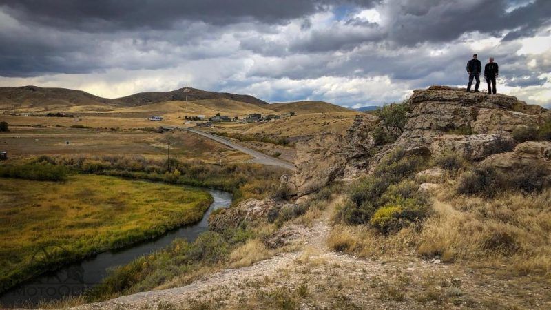 Two hikers on a rocky hill overlook a winding stream and cloudy open moorland