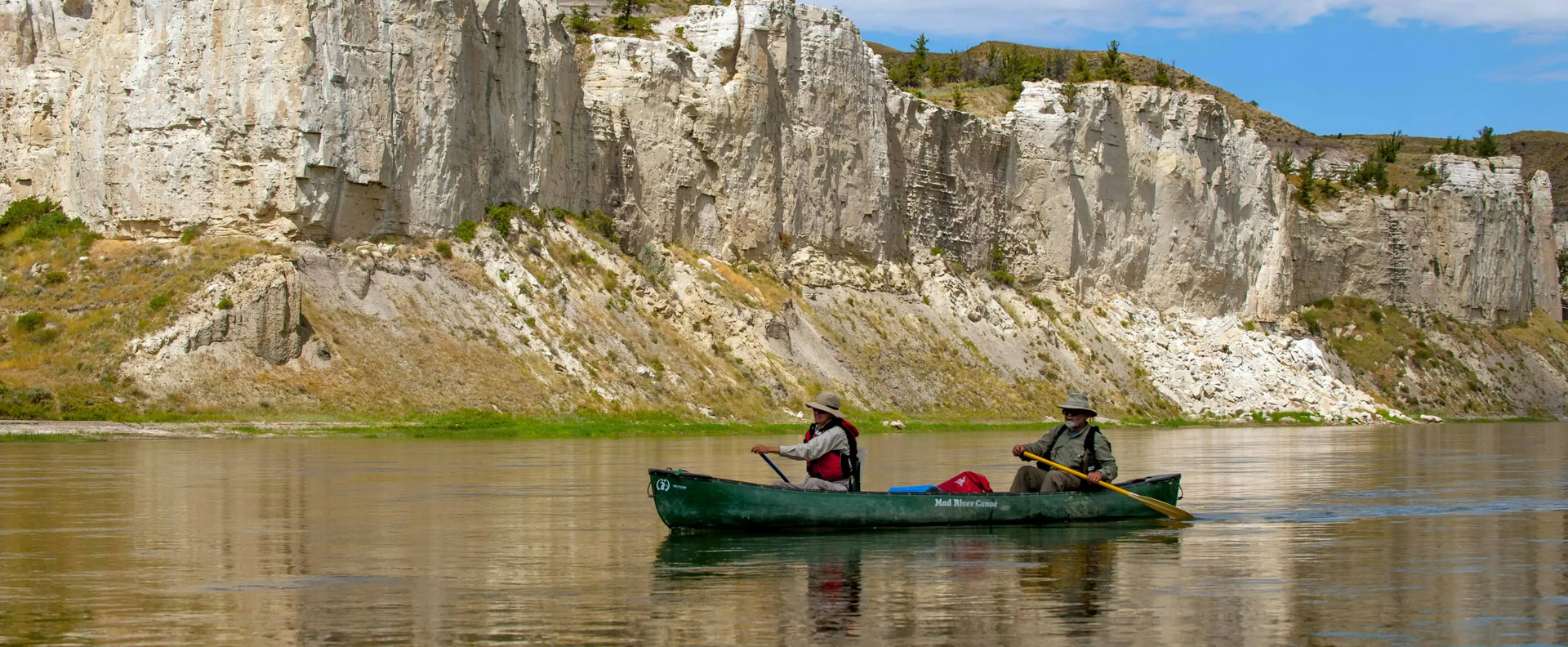 Two people paddle a green canoe on calm water before a tall rocky cliff.