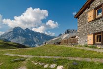 Stone houses in a mountain village beside a grassy meadow and snow-capped peak under a blue sky