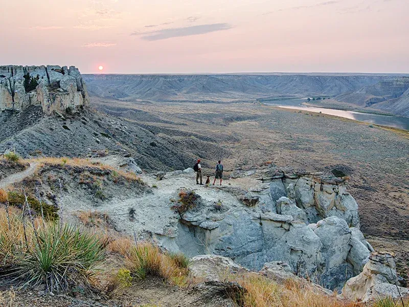 Sunset over a rocky canyon with two hikers on a cliff above a winding river