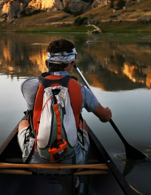 Person kayaking on a calm river at sunset, seen from behind, with red backpack and mountain reflections.