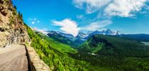 Mountain road overlooking a green valley and snow-capped peaks under a blue sky