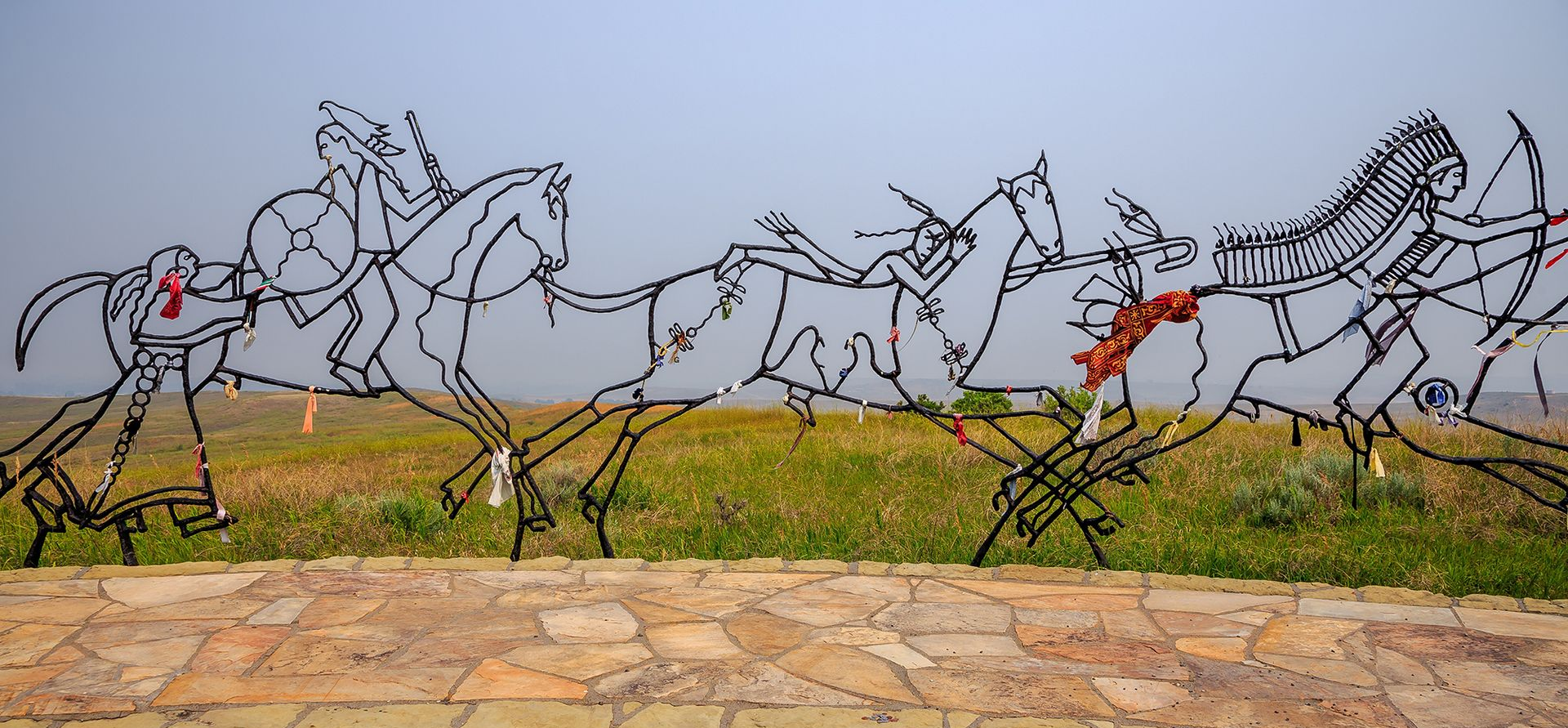 Metal horse sculptures in motion along a grassy roadside with stone pavement in front.
