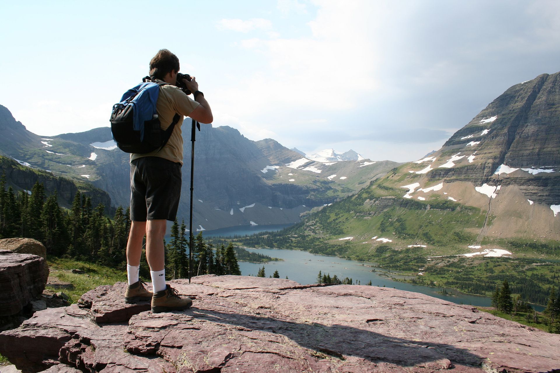 Hiker with backpack taking a photo from a rocky overlook above a mountain valley and lake