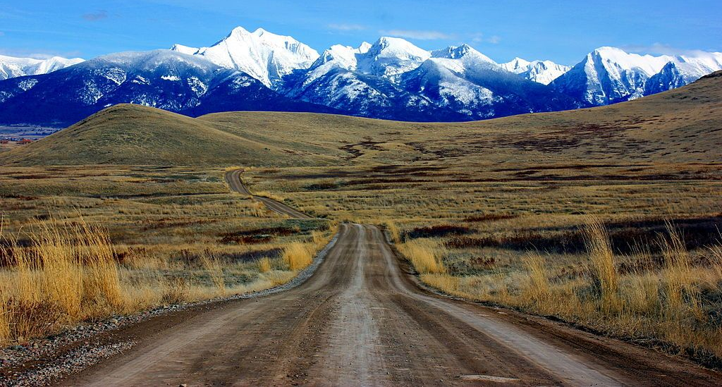 Dirt road leading through a grassy plain toward snowcapped mountains under a clear sky