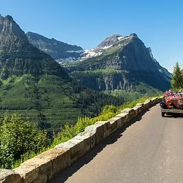 Mountain road beside a stone guardrail, overlooking green cliffs and snowy peaks under a clear blue sky