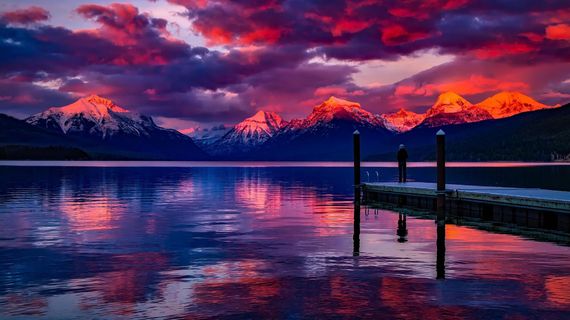 Pink sunset over snowy mountains reflected in a calm lake, with a dock in the foreground.