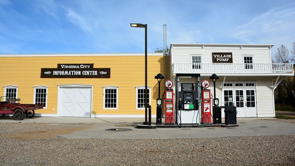 Old western-style storefronts with a yellow building, white building, and vintage gas pumps along a gravel road