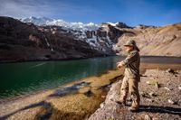 Person fishing beside a turquoise mountain lake with snow-covered peaks in the background