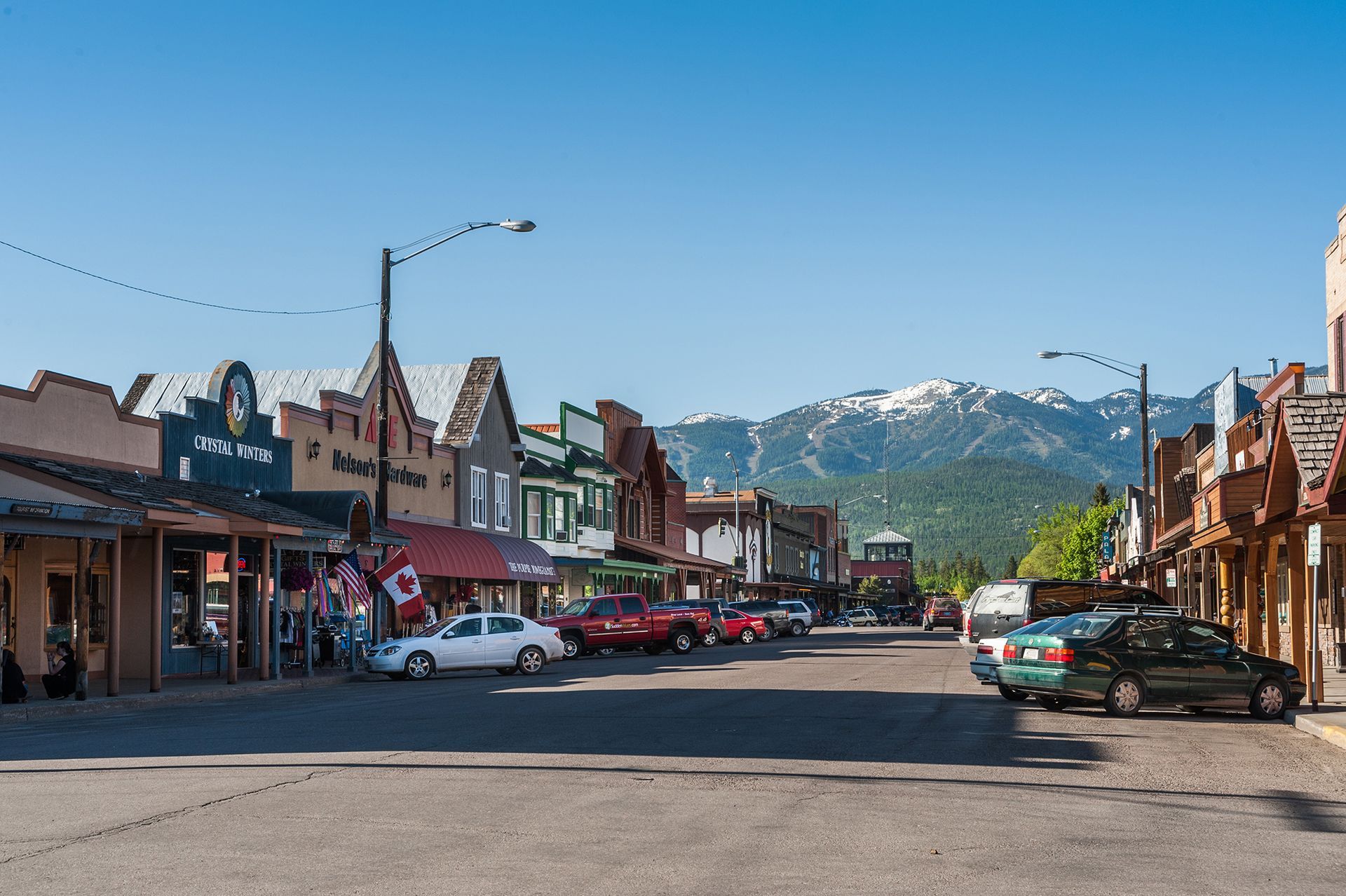 Small-town main street with parked cars, storefronts, and snowy mountains in the background