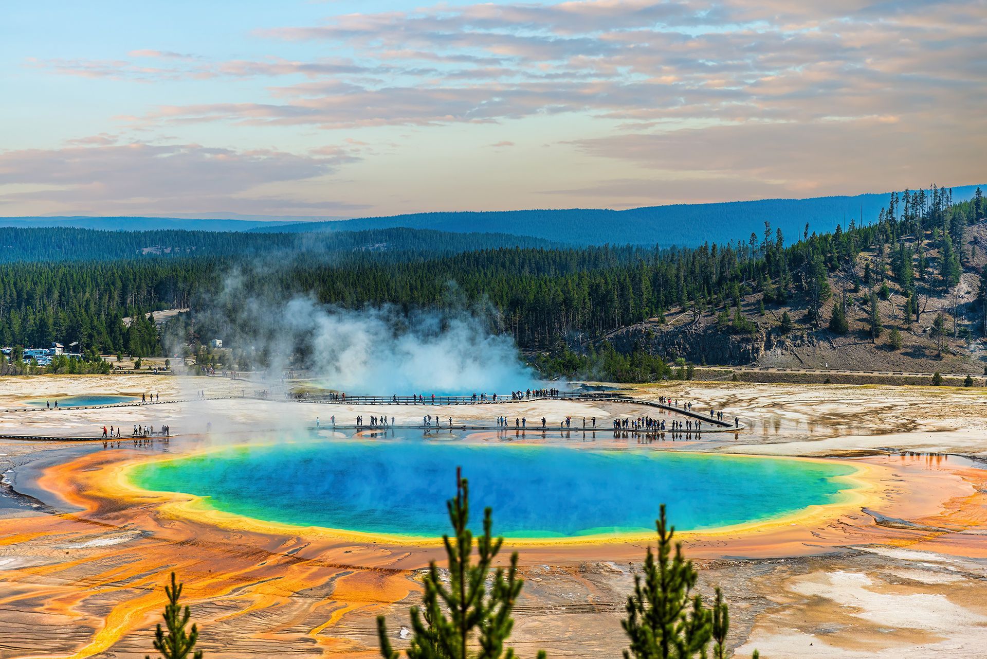 Rainbow-colored hot spring pool steaming in a geothermal landscape with forest and sky beyond