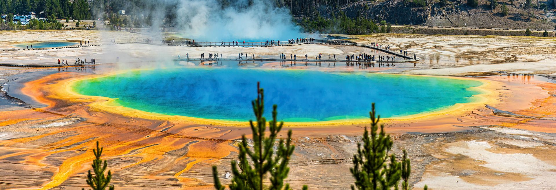 Colorful geothermal hot spring with a turquoise center, orange mineral edges, and steam rising from the water