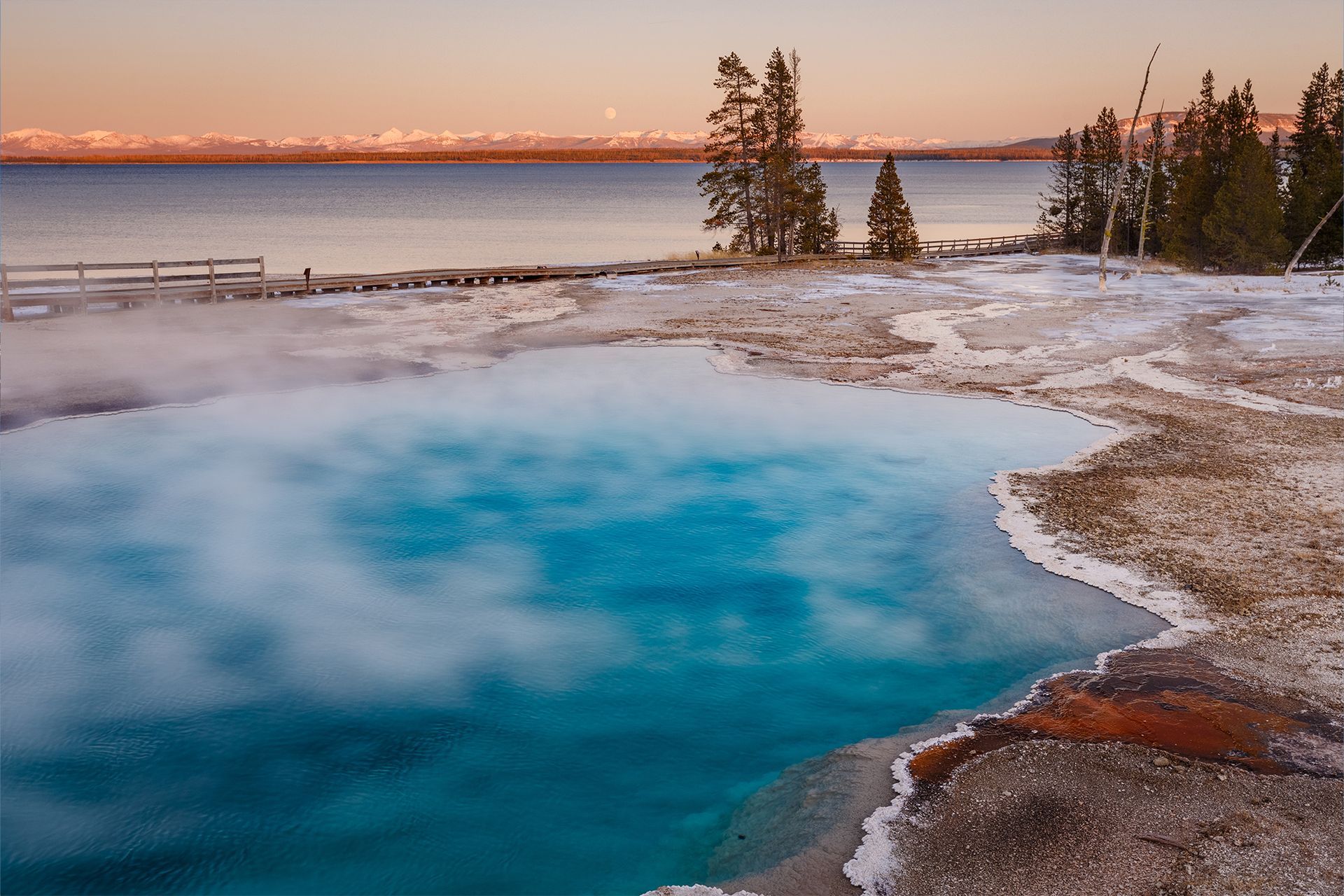 Steaming blue hot spring beside a snowy shoreline at sunset, with trees and a calm lake in the background