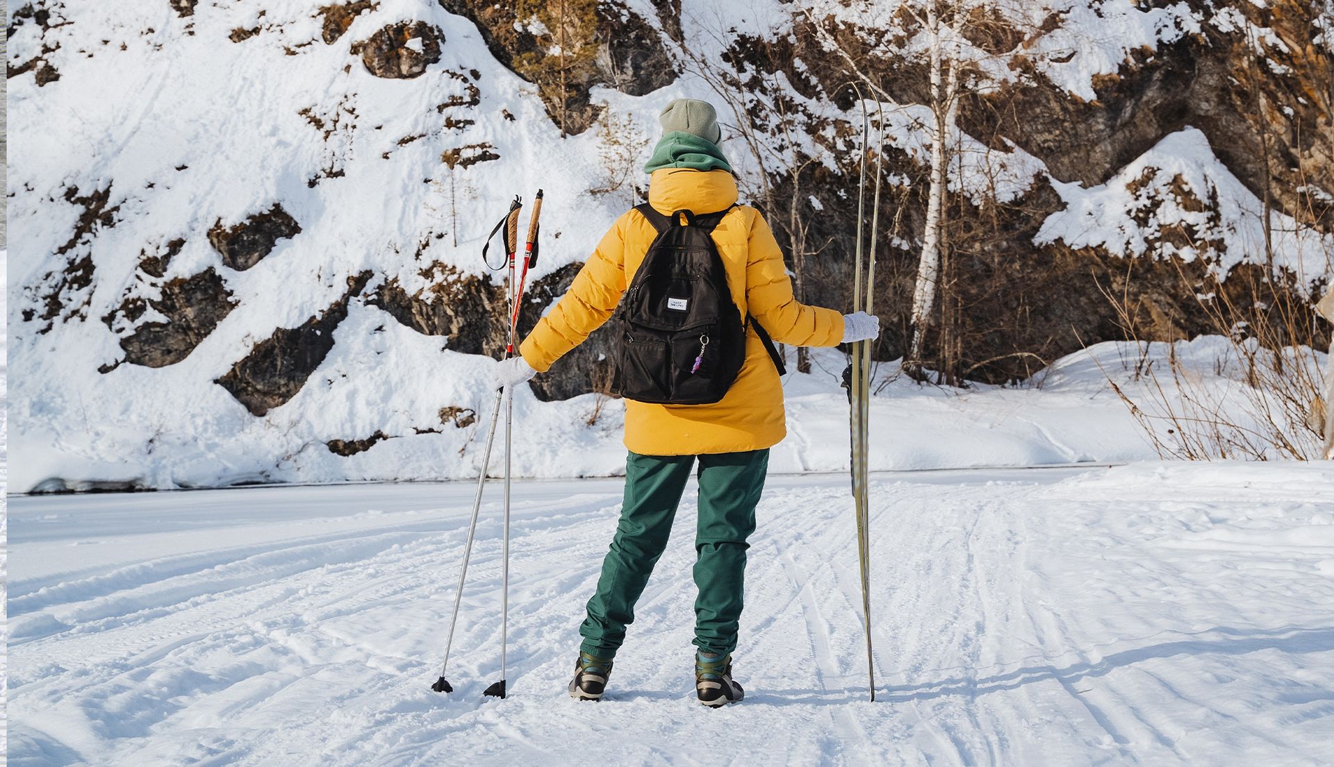 Skier in yellow jacket and green pants stands on snowy trail with poles near rocky hillside