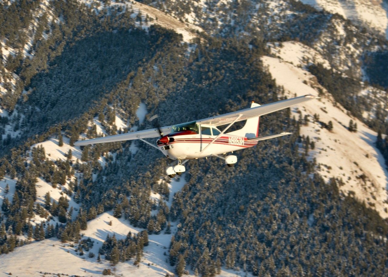 White-and-red small airplane flying over snowy forested mountains.