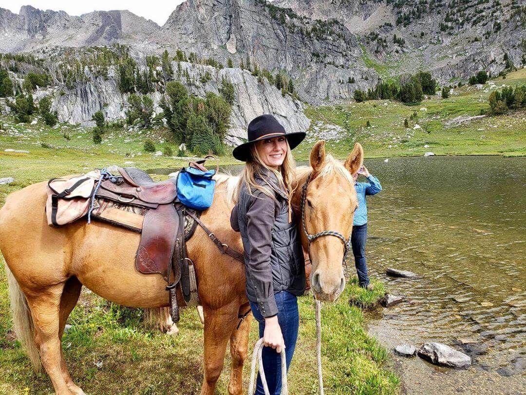 Woman in black hat stands beside saddled horse by a mountain lake with rocky cliffs in the background