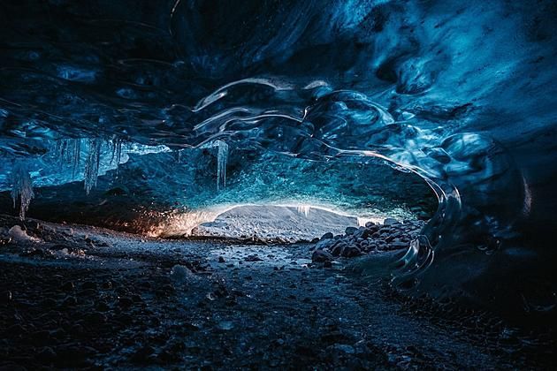 Blue ice cave with a glowing opening and a dark, rocky floor