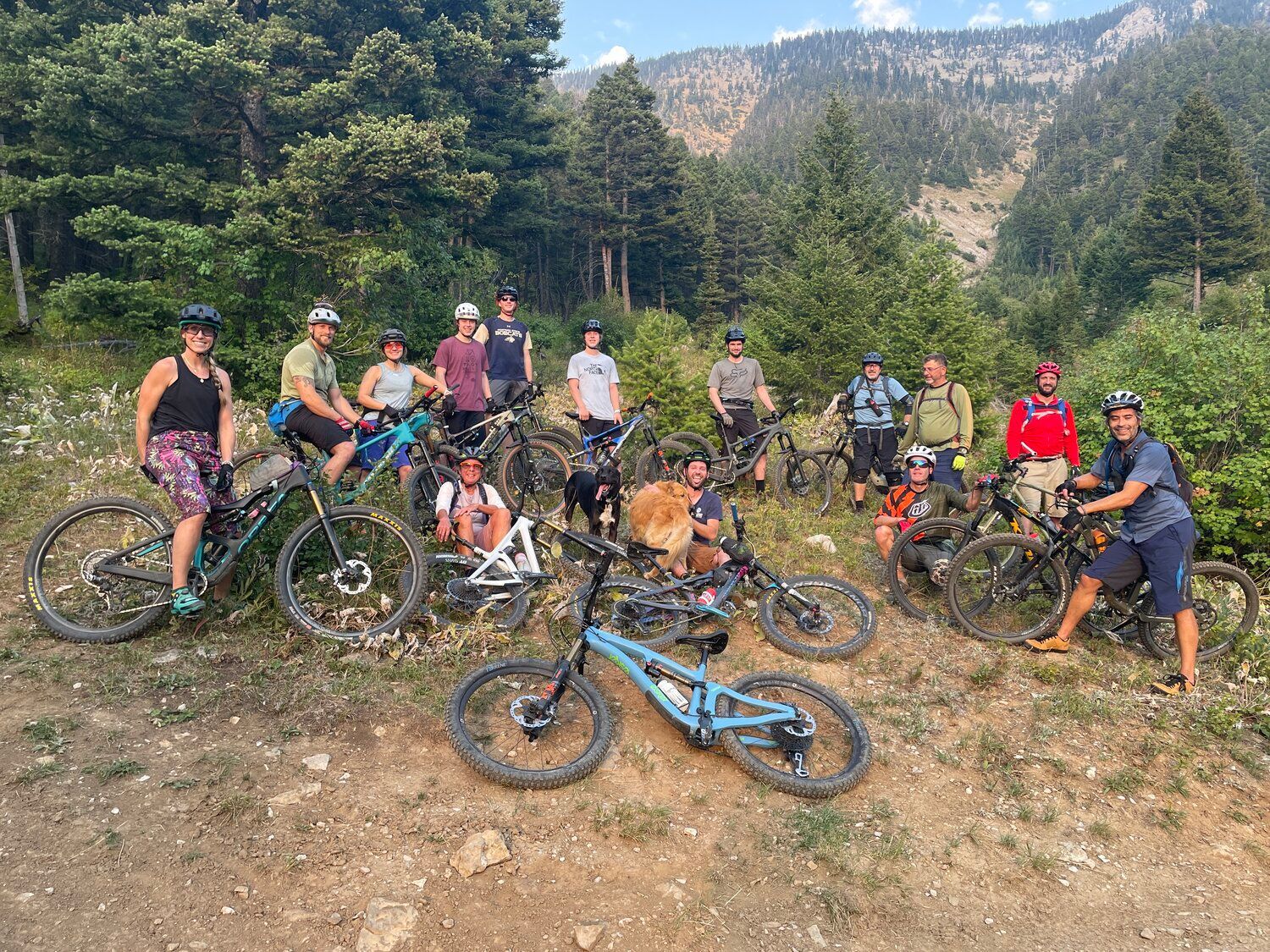 Group of mountain bikers posing with their bikes on a dirt trail in a wooded mountain area