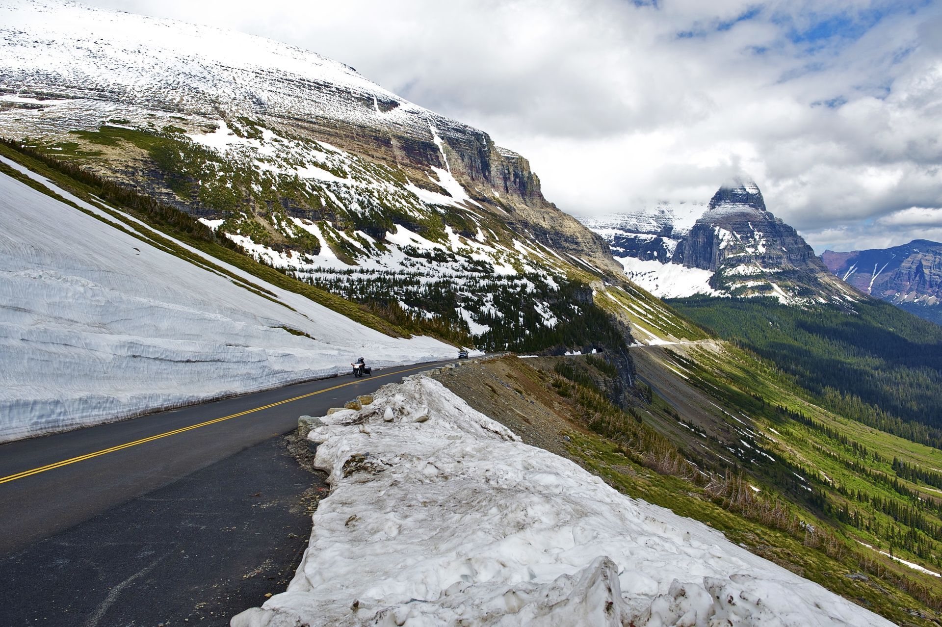 Mountain road with snowbanks overlooking a green valley and snow-capped peaks under cloudy skies