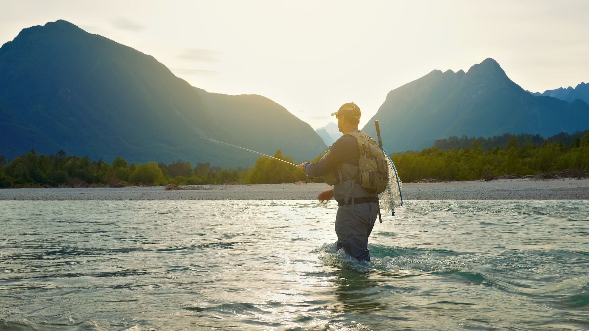 Person wades through a sunlit river with mountains silhouetted in the background.