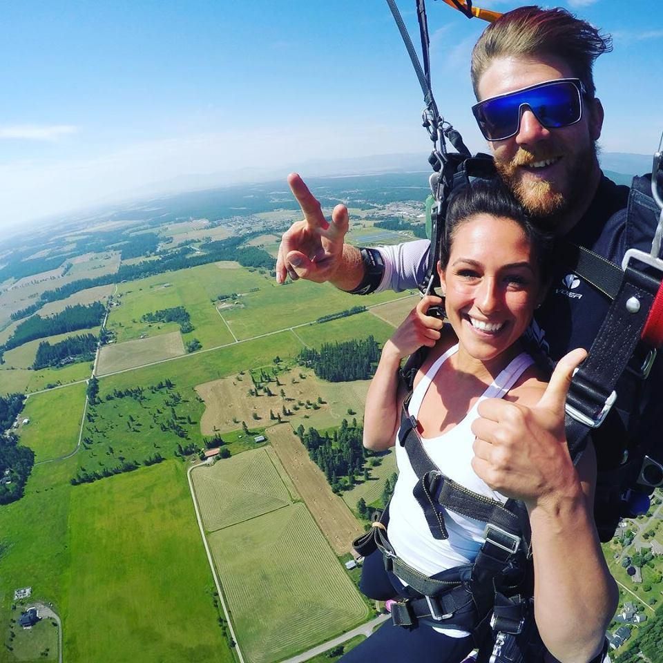 Two smiling skydivers in harnesses, one giving a thumbs-up above a green farmland landscape.