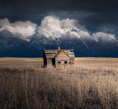 Abandoned wooden cabin in a dry field beneath dark storm clouds and snowy mountains