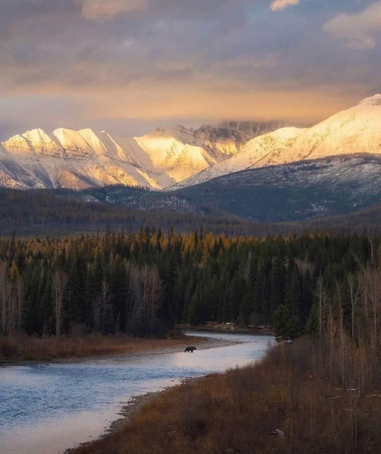 Snowy mountain range above a winding river and dark forest at sunset.